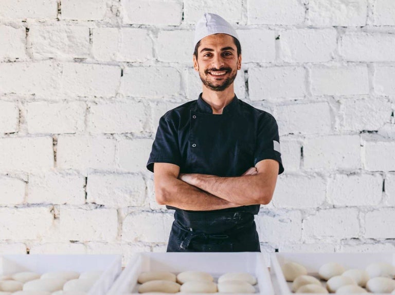 Emprendedor gastronómico sonriendo con los brazos cruzados frente a cestas de masas para panadería