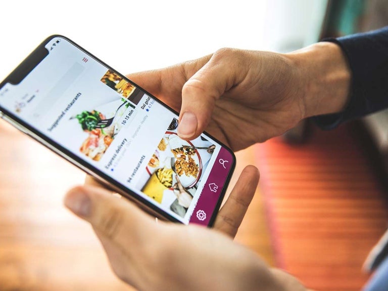 Manos sostienen un celular que muestra una plataforma de entrega a domicilio con diferentes platillos