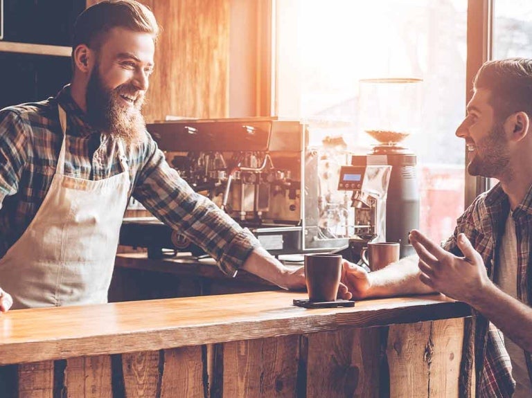 Barista conversa animadamente con cliente sentado a la barra de su negocio que sostiene una taza de café
