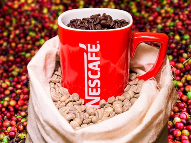 Taza roja llena de café en grano NESCAFÉ® en una bolsa con café verde y rodeado de cerezas