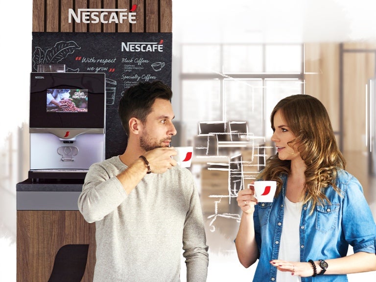 Dos compañeros de trabajo disfrutan de una taza de café en la oficina, al fondo una máquina NESCAFÉ®