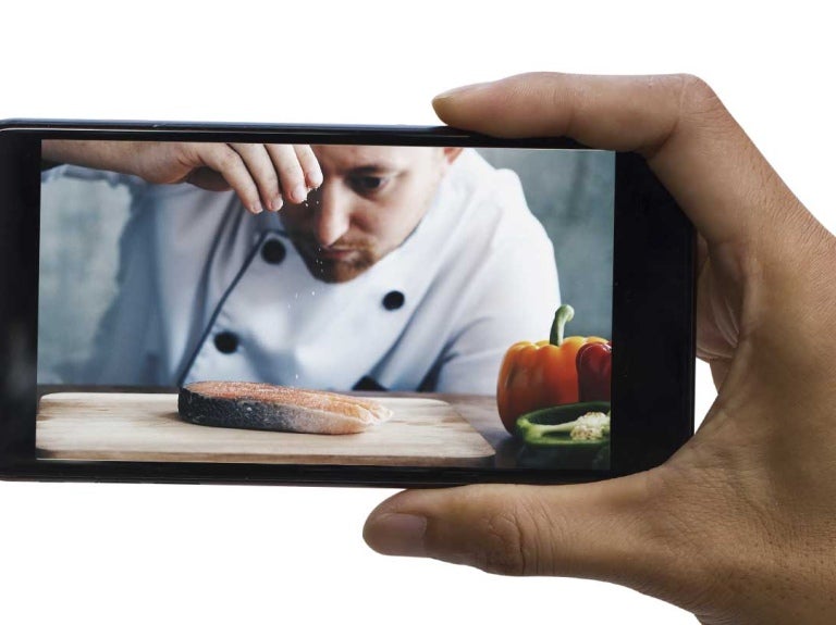 Mano sostiene un smartphone en el que se proyecta un chef sirviendo diferentes platillos en una cocina