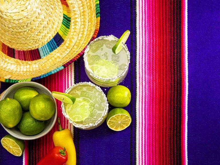 Dos vasos de michelada con rodajas de limón en el borde y al lado la fruta, un sombrero y maracas