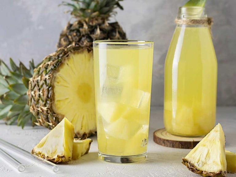 Vaso y botella de cristal de agua saborizada de piña, al fondo está la fruta y los trozos