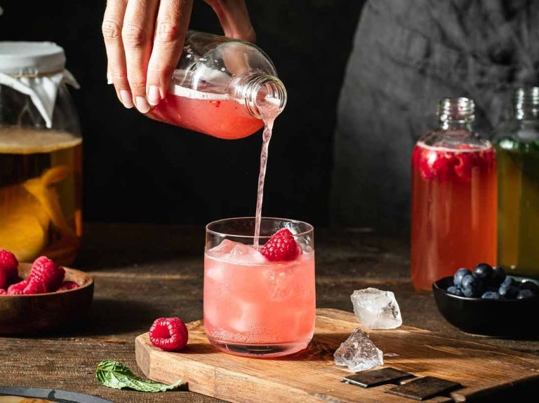 En vaso con hielo y frambuesas mixólogo vierte un líquido de una botella, al lado otras bebidas y frutas