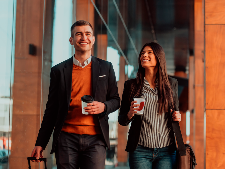 Hombre y mujer sonrientes, caminando en aeropuerto con bebidas de NESCAFÉ en mano y equipaje
