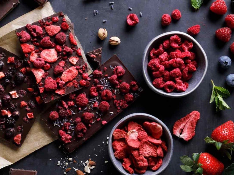 Tabletas de chocolate con frambuesas, arándanos y fresas liofilizadas, al lado las frutas frescas