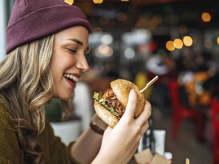 Mujer observa una hamburguesa que sostiene en sus manos, al lado bebida y otros aperitivos
