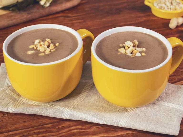 Dos tazas amarillas de chocolate caliente coronadas con maní, al fondo hallacas sobre una tabla