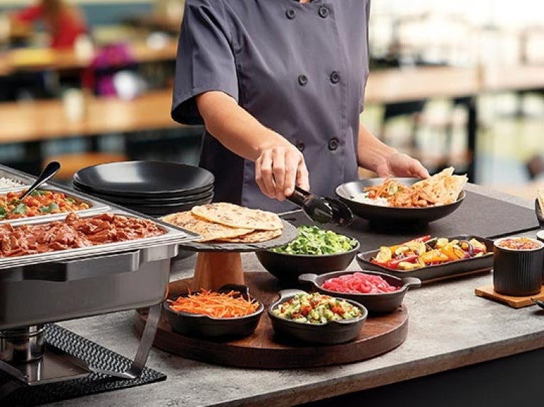 Chef frente de una mesa de buffet sostiene unas pizas para tomar la ensalada de una taza