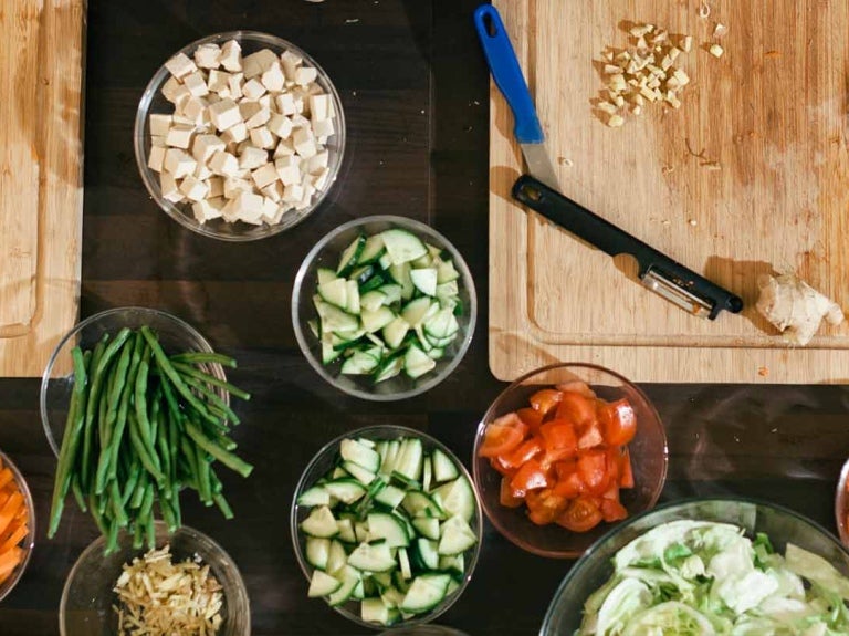 Mesa con tablas de cortar, tazas con alimentos cortados de distintas formas, alrededor, manos de mujeres