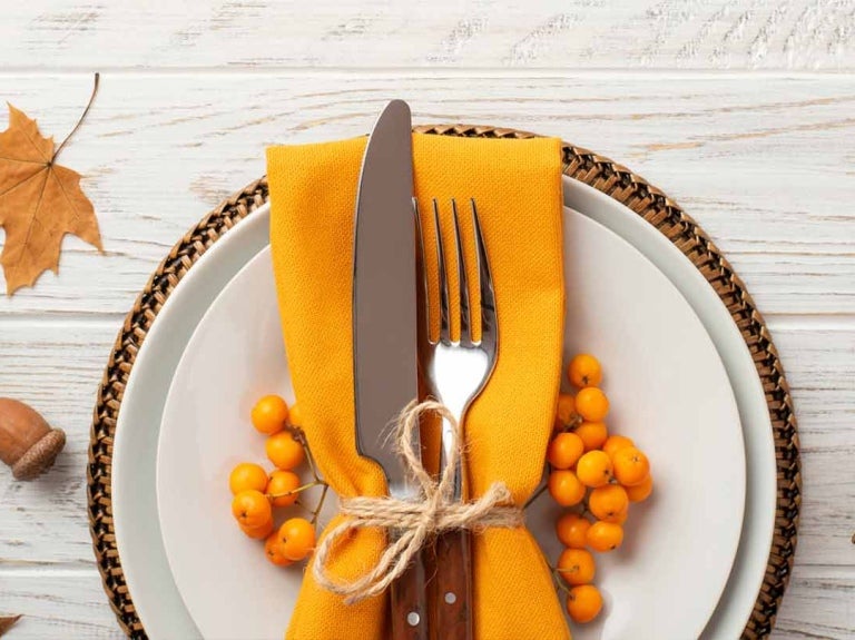 Tenedor y cuchillo atados a una servilleta naranja sobre un plato, alrededor decoraciones de otoño