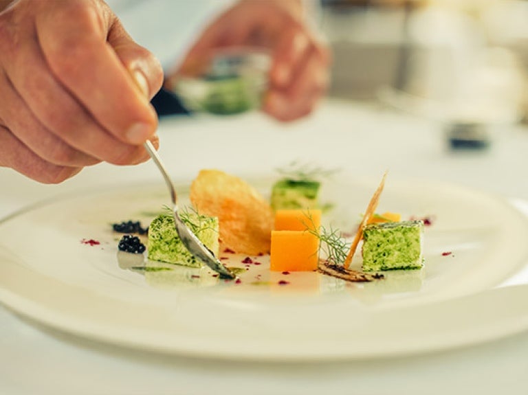 Mano de chef decora con aderezo un plato blanco de vegetales en cubos