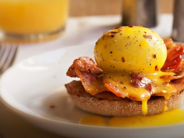 Huevo benedictino con bacon y salsa holandesa sobre un muffin inglés, al fondo jugo y saleros
