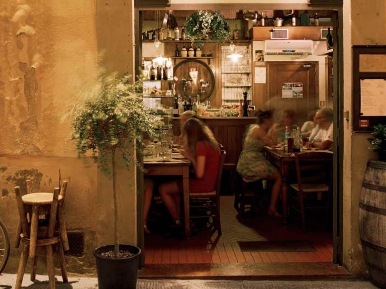 Fachada de una hostería con comensales al fondo sentados a la mesa y afuera sillas, plantas y bicicleta
