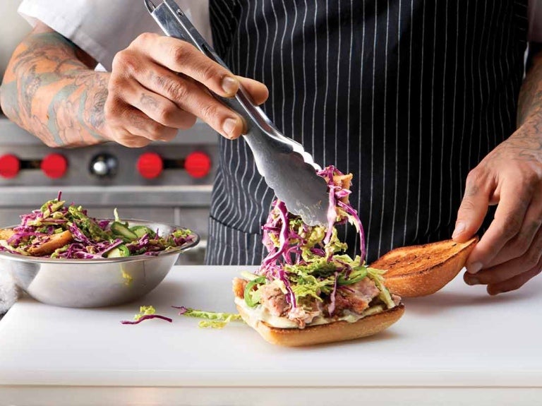 En una cocina comercial, chef agrega ensalada con pinzas al pan sobre una tabla de cortar