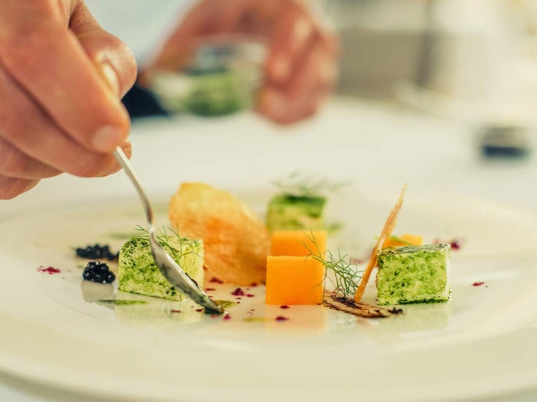 Mano de chef decora con aderezo un plato blanco de vegetales en cubos