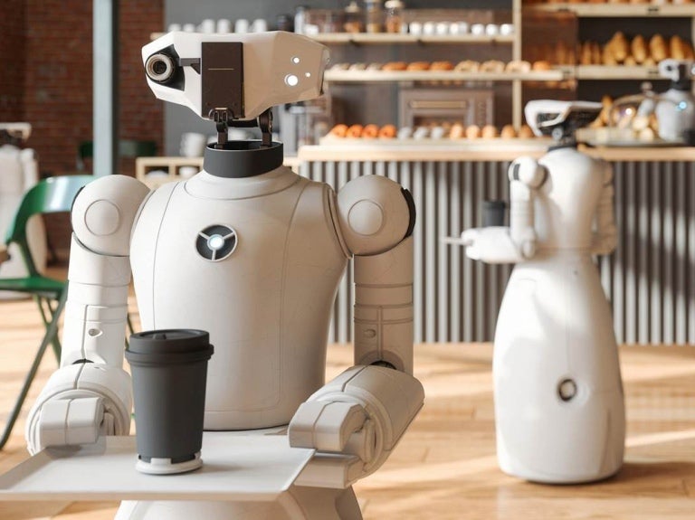 Dos robots recorren una cafetería sosteniendo una bandeja con una bebida caliente con tapa
