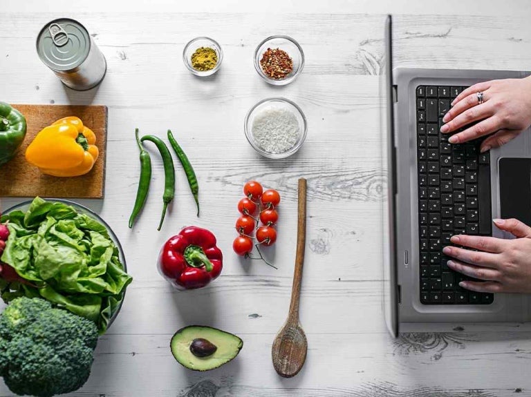Ingredientes frescos como vegetales, especias y arroz frente a una laptop usada por las manos de una mujer