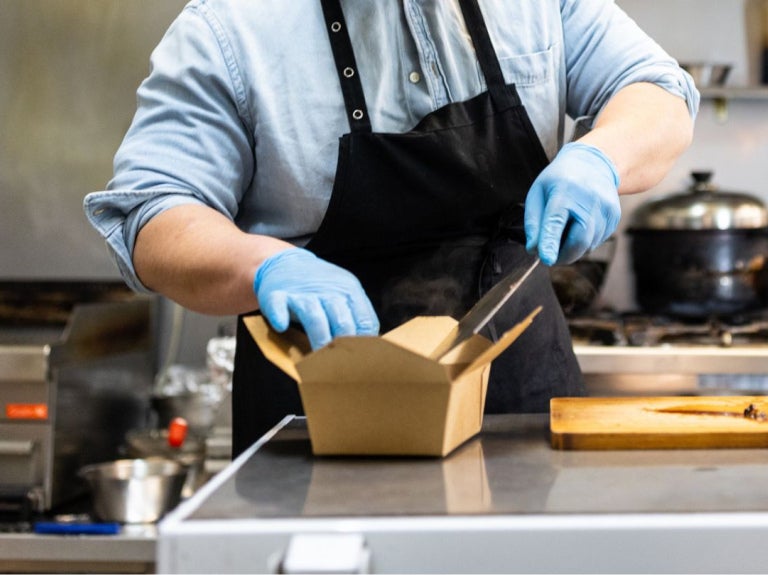 Chef con guantes y delantal empacando comida en una caja para llevar dentro de una dark kitchen profesional