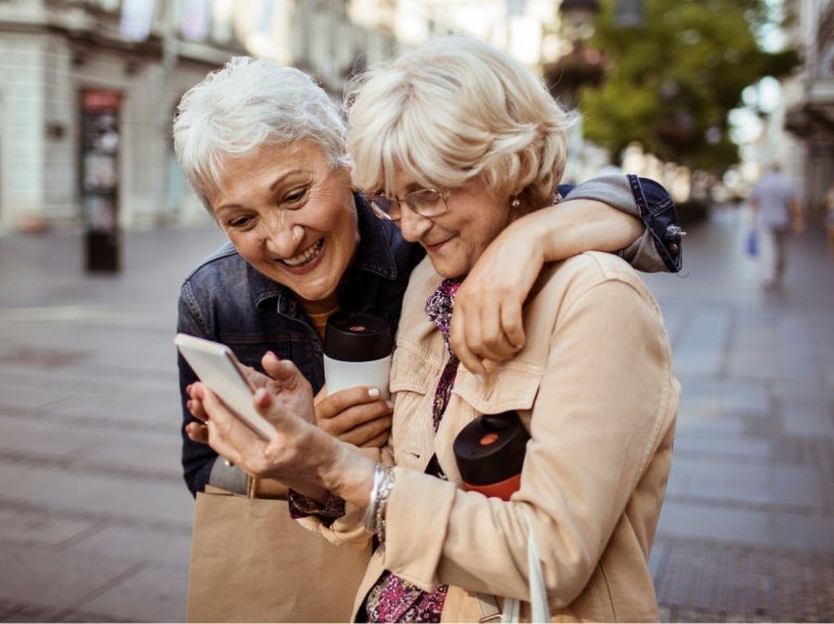 Dos mujeres mayores sonrientes observan un celular mientras pasean por una ciudad con café en mano