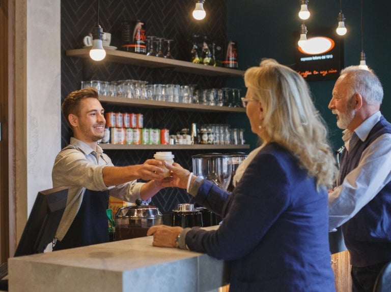 Barista entrega una bebida a una clienta en cafetería mientras otro hombre espera