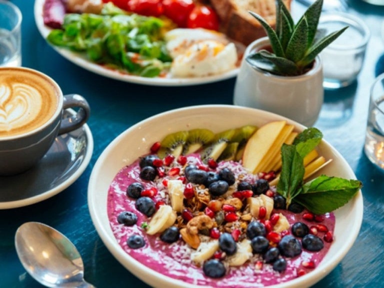 Smoothie bowl decorado con frutas frescas, acompañado de un café latte y un desayuno variado en una mesa.