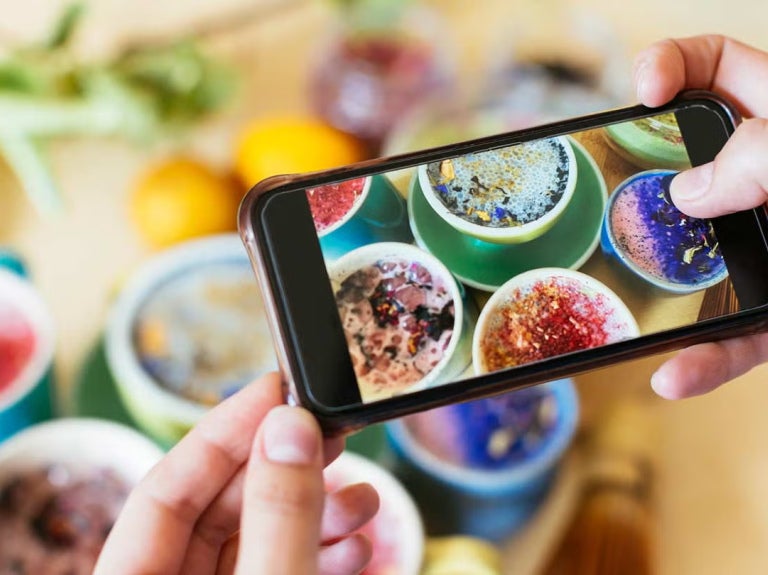 Persona tomando una foto con el celular a varias tazas coloridas de bebidas decoradas con flores y semillas