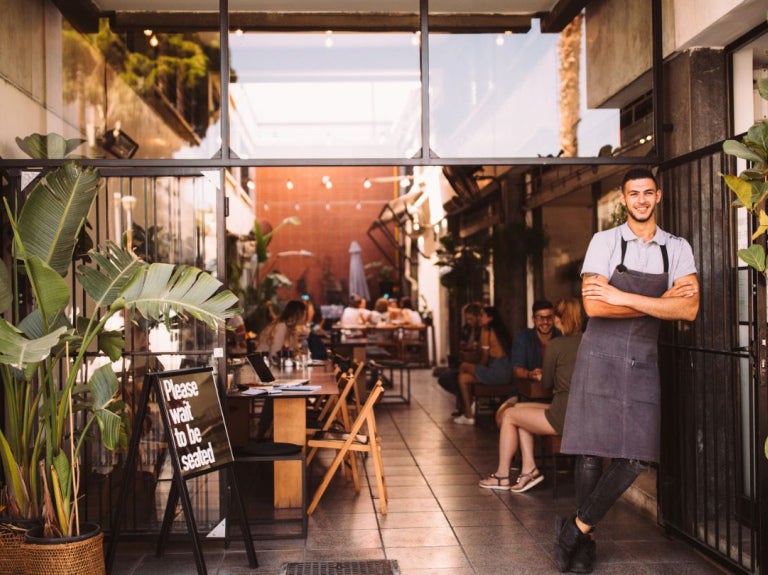 Entrada de un restaurante con plantas y mesas al aire libre, creando un ambiente tipo jardín gastronómico.