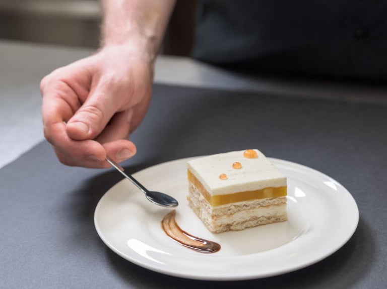 Mano de chef pastelero sostiene una cuchara con la que decora el plato de un postre con salsa