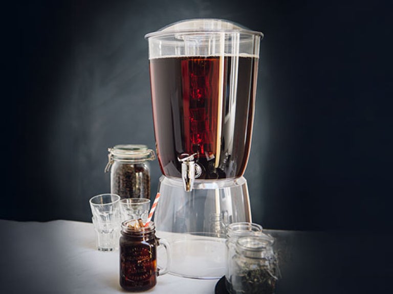 Dispensador de café cold brew junto a vasos y frascos con granos de café en una mesa.