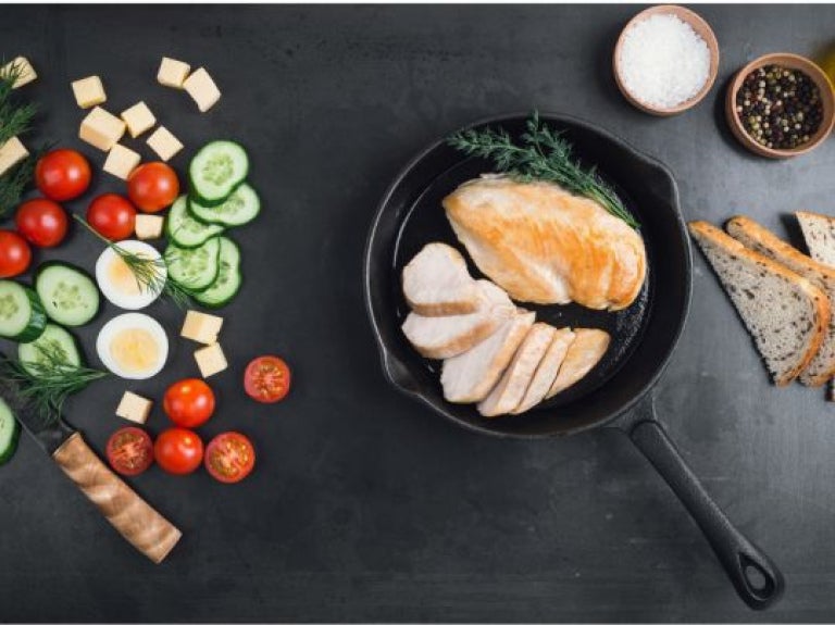 Sartén con pollo rodeado de verduras frescas, pan y especias sobre una mesa negra