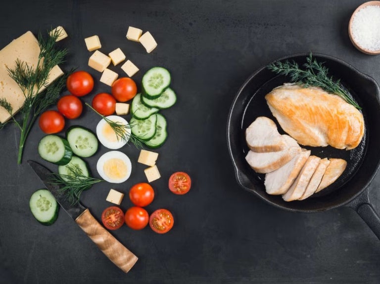 Sartén con pollo rodeado de verduras frescas, pan y especias sobre una mesa negra