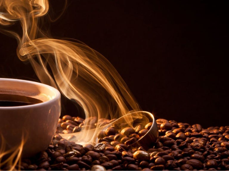 Taza de café humeante entre granos tostados, con el vapor formando curvas en un ambiente oscuro