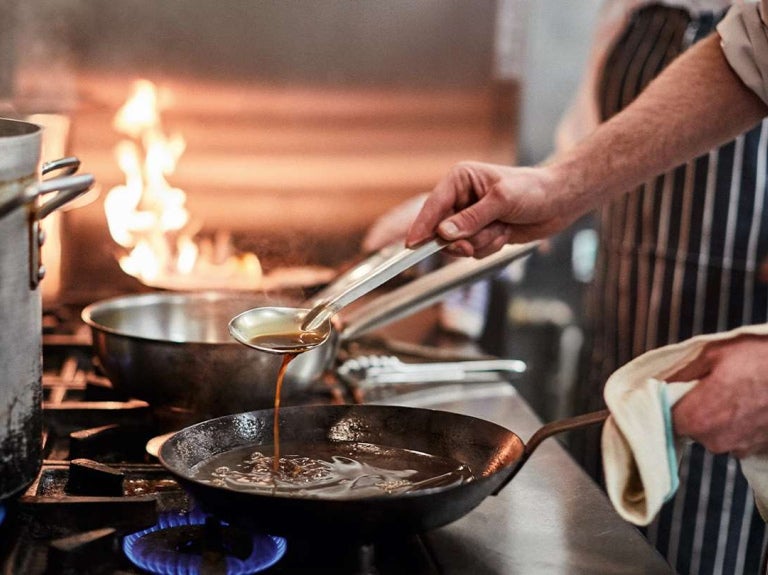 Chef vertiendo salsa en sartén caliente dentro de una cocina profesional en plena preparación
