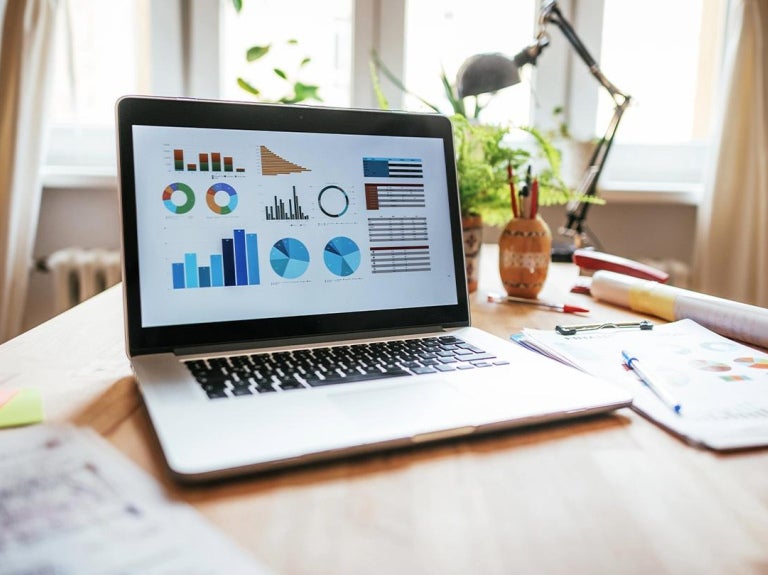 Escritorio con portátil mostrando gráficos y datos, rodeado de documentos, útiles y plantas