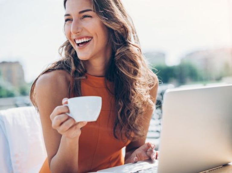 Mujer sonriente sosteniendo taza frente a portátil en mesa exterior