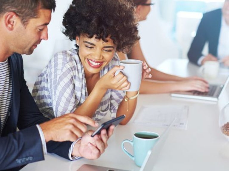 Personas en oficina compartiendo café mientras revisan celular y trabajan en portátil