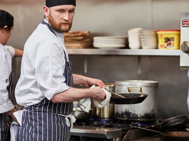 Chef cocinando con sartén frente a estufa en cocina profesional con otros cocineros