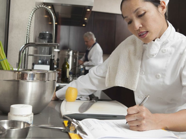 Chef escribiendo en libreta en cocina con utensilios y alimentos sobre mesa