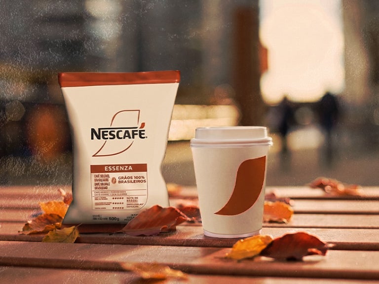 Bolsa de café Alegría Espresso NESCAFÉ® y vaso desechable sobre mesa con fondo urbano desenfocado
