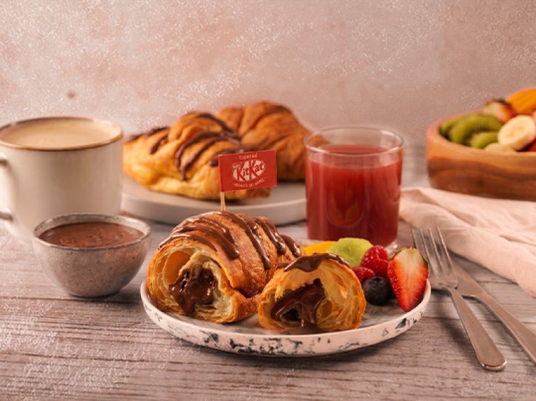 Croissant relleno con chocolate KITKAT® acompañado de frutas, bebida y café en mesa