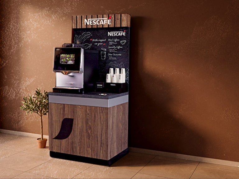 Máquina NESCAFÉ® instalada sobre módulo con vasos al costado en el lobby de un hotel