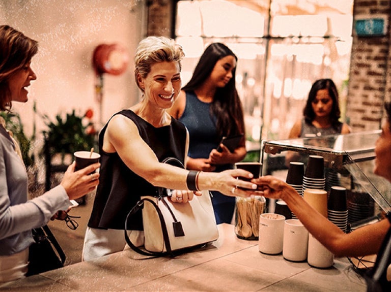 Cliente recibe vaso de café en mostrador en una cafetería con una fila de clientes