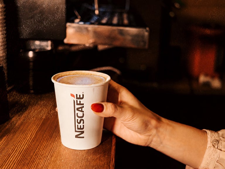 Mano sosteniendo un vaso de NESCAFÉ® servido sobre barra de preparación en una cafetería