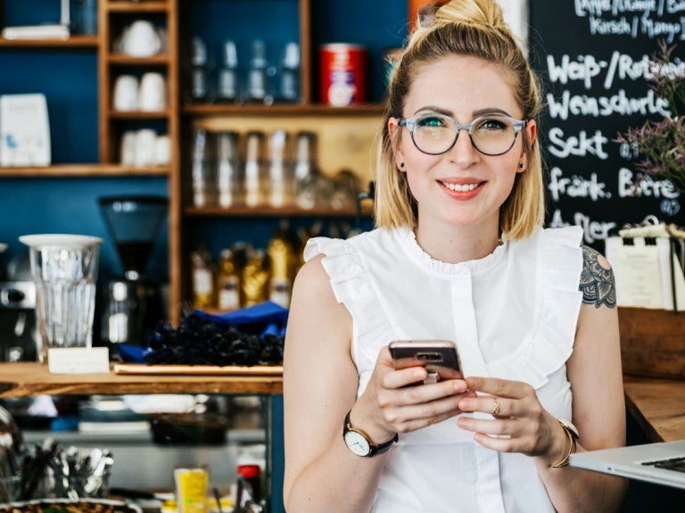 Mujer en cafetería usando celular y laptop, gestionando tareas en entorno de negocio.