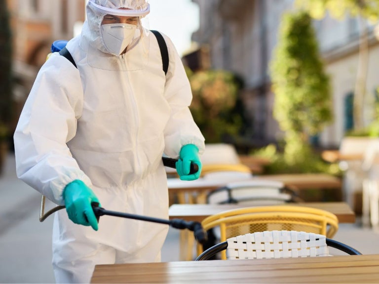Técnico con traje de protección fumiga en la zona de las mesas de un restaurante al aire libre.