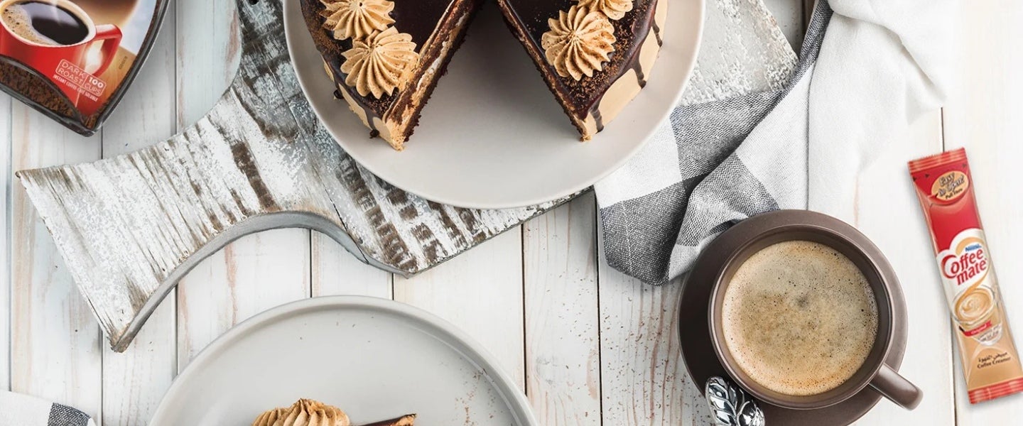 Una rebanada de pastel de chocolate y alrededor un frasco de café y crema para café de un proveedor al por mayor.