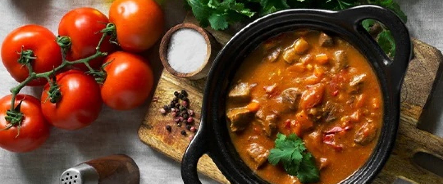 Olla de estofado rodeada por tomates, aceite, hierbas aromáticas y otros productos culinarios
