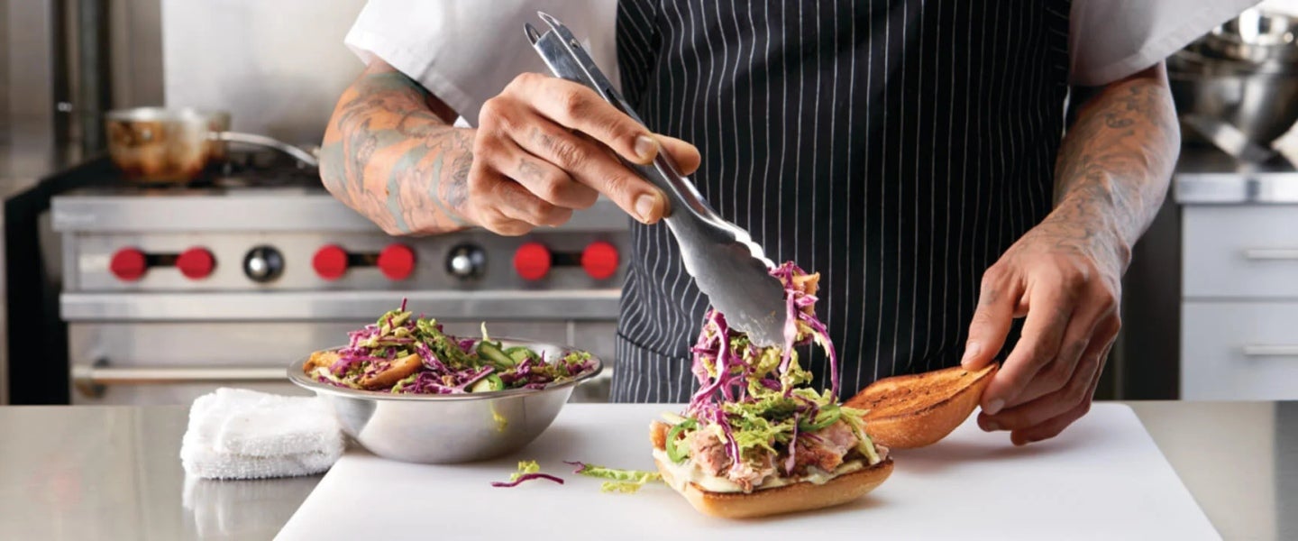 Manos de chef agrega una ensalada de una taza utilizando unas pinzas, en una cocina comercial